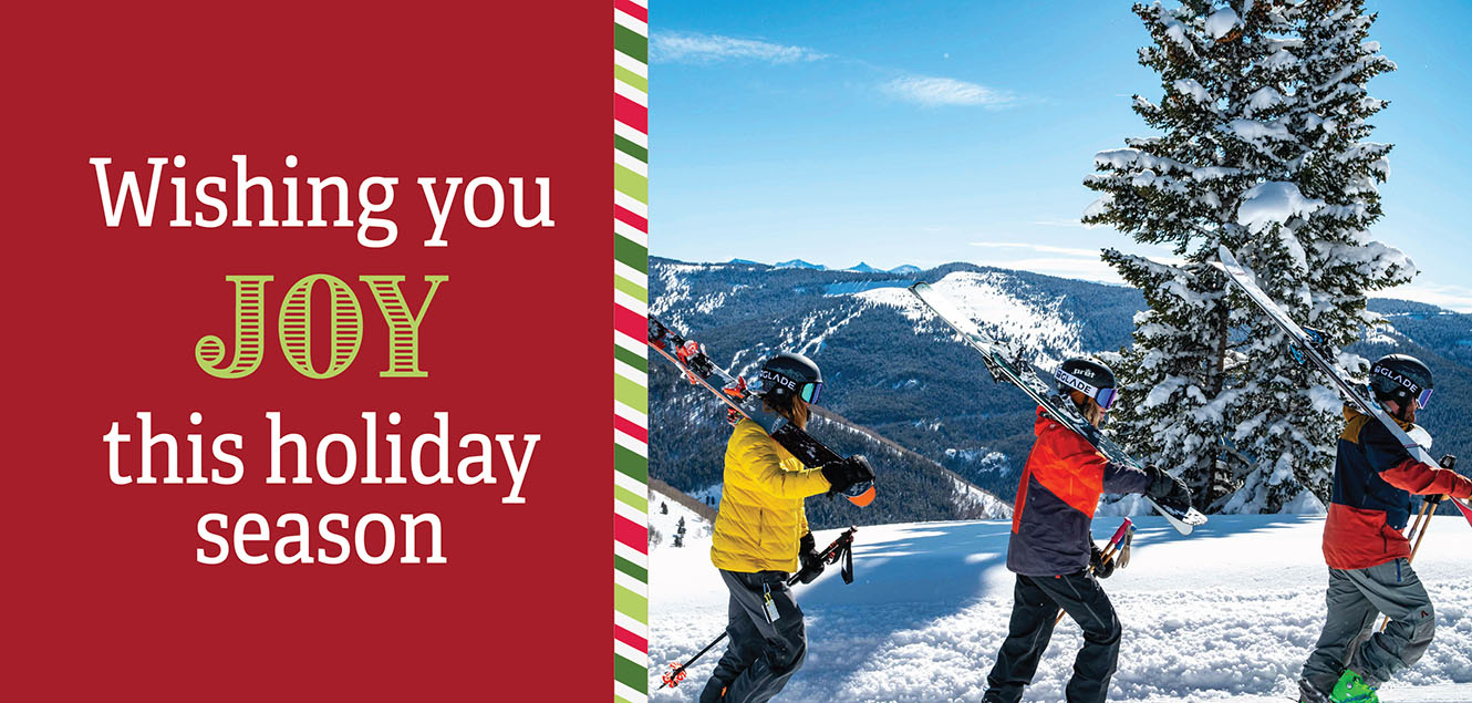 wishing you joy this holiday season with three people carrying skis through the snow